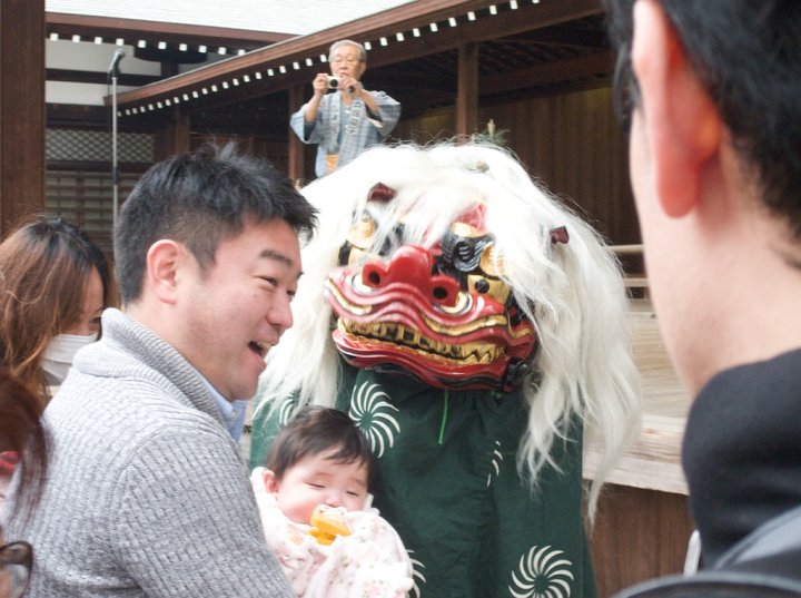 A man raises up his baby girl to the dancing lion. The lion bites her head, then that of her dad. As a result they both can expect good luck for the coming year. This is a lion dance at Yasukuni Shine, Tokyo, Japan.