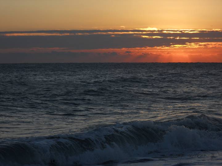 The morning sun sets the clouded sky on fire above the dark and churning white-tipped blue of the Atlantic Ocean shoreline near Melbourne Beach, Florida. The condensed water particles in the lower atmosphere over the ocean break the sun into colors. The red color results from reflection and refraction of sun light passing through the atmosphere and touching the clouds. Snapped with a Nikon Coolpix L22. Copyright 2010 by Village Hiker Publishing Company. All rights reserved. Please do not use without permission. Contact Village Hiker for digital reproduction and high-resolution prints.