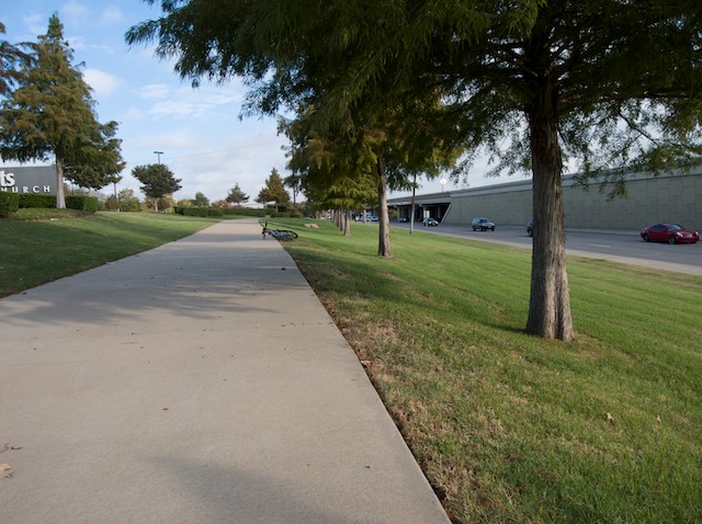 Vehicles move southbound along the North Central Expressway access road. While the hiking and biking trail continues along a line of cedar trees you have exited from the Spring Creek Nature Area.