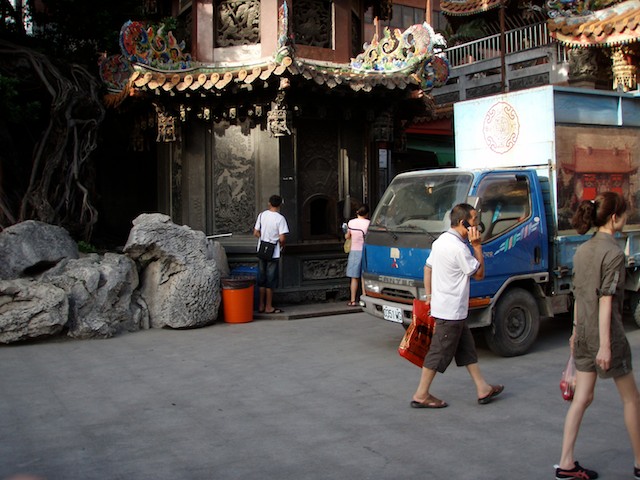 A man with a cell phone enters the temple, while a woman in the background burns temple money as a gift to ancestors. This is temple for Matsu in the Dajia District of Taichung, Taiwan.